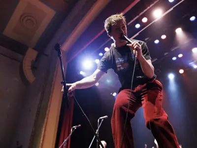 A musician sings into a handheld microphone on stage under bright concert lights, leaning toward the audience while performing live at a theatre.